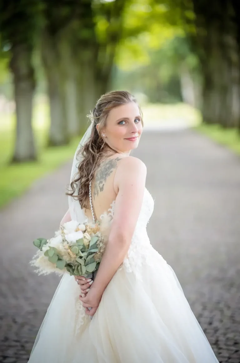 Braut mit Hochzeitskleid und Blumenstrauß, auf einem schmalen Weg zwischen Bäumen.
