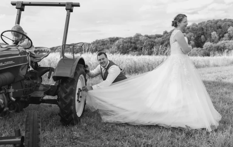 Heiraten in Bad Driburg Braut mit schwingendem Kleid und Mann, der an einem Traktor arbeitet, im Freien.