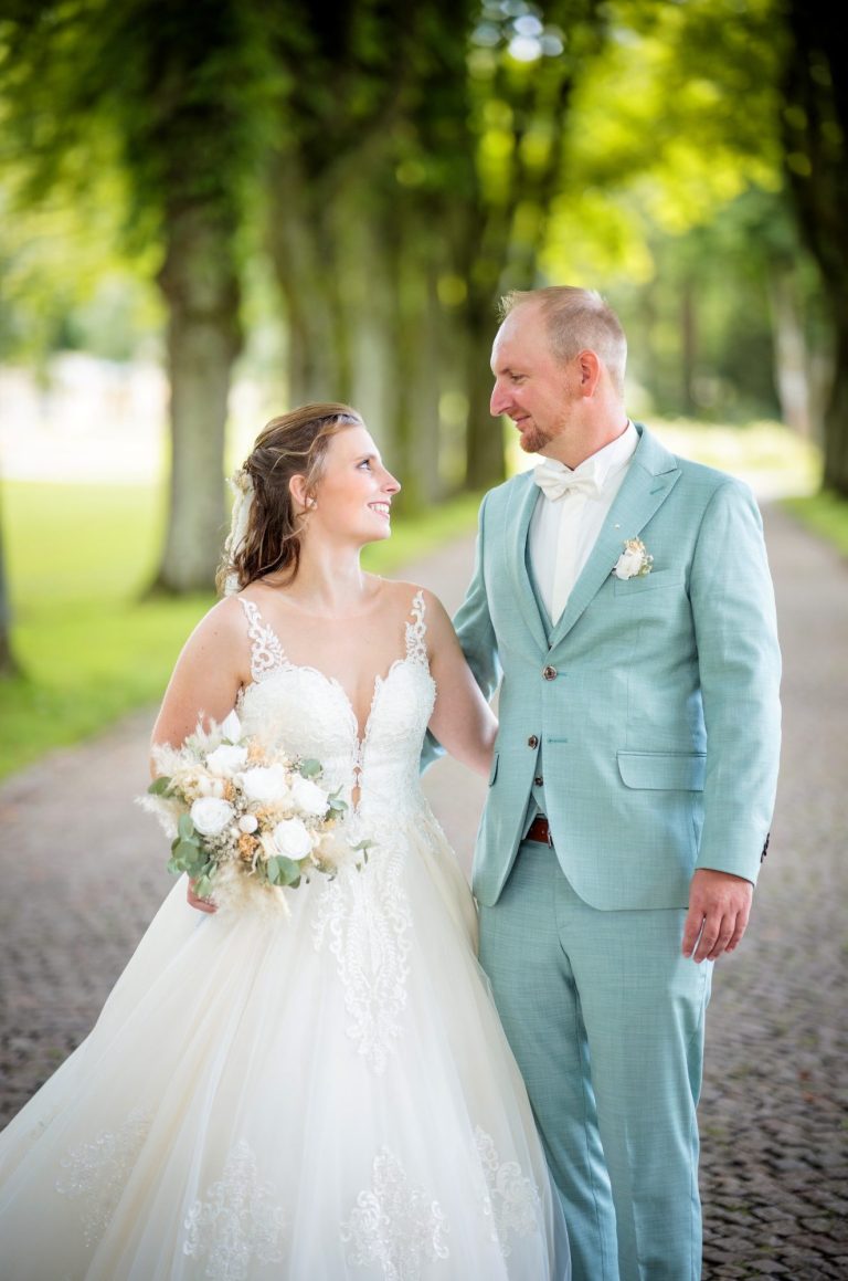 Braut und Bräutigam in eleganter Hochzeitskleidung, umgeben von Bäumen auf einem Weg.