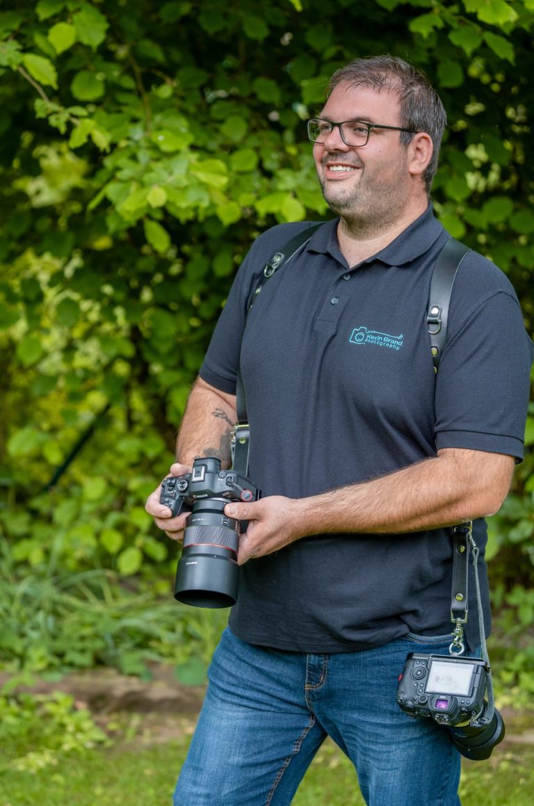 Hochzeitsfotograf aus Bad Driburg im Kreis höxter Ein lachender Hochzeitsotograf hält eine Kamera und steht in einem grünen Garten.