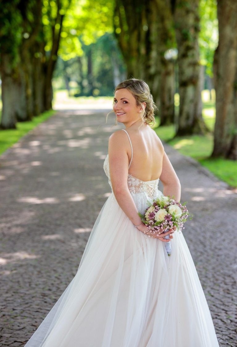 Braut aus Höxter Braut in einem eleganten Kleid, steht auf einem Baum gesäumten Weg und hält einen Blumenstrauß.